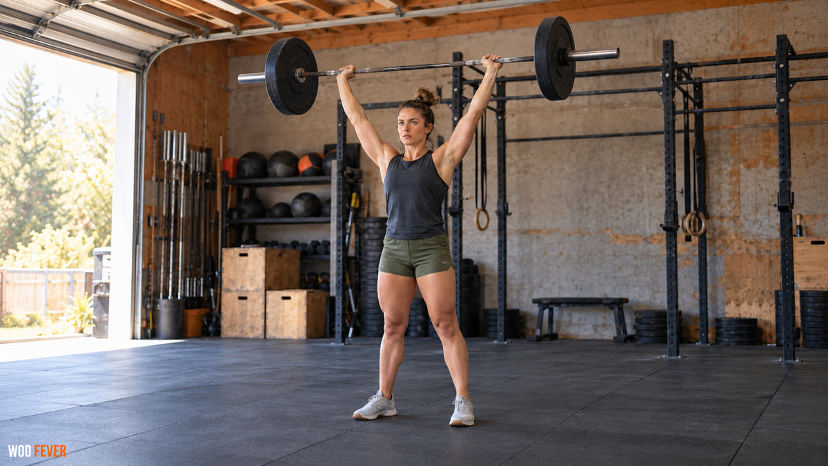 Athlete at the overhead finish of a barbell thruster with a pull-up rig behind her for the Fran workout.